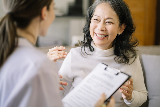 Woman has her back towards the camera  holding a clipboard and is speaking to a middle-aged asian woman that is smiling.